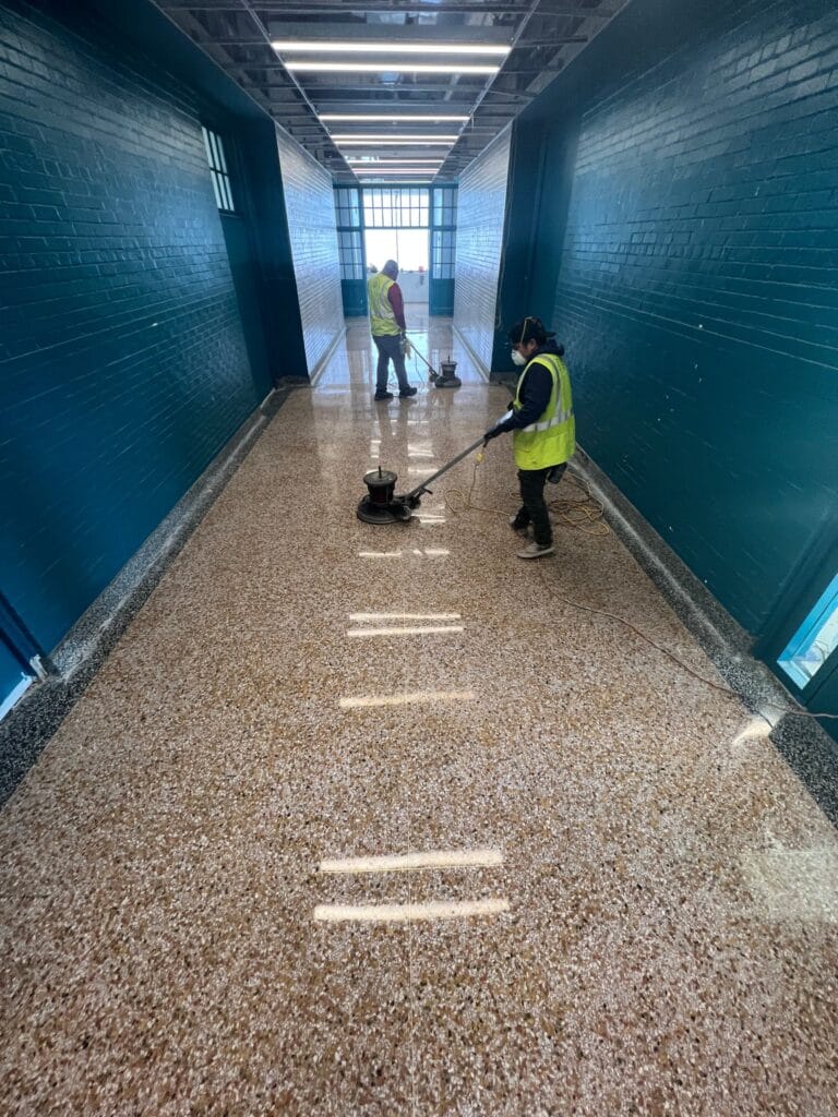 Two technicians in high-visibility vests polishing a terrazzo school corridor with a floor machine